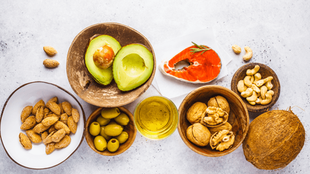 Assorted healthy high-fat foods such as avocado, salmon, almonds, walnuts, cashews, green olives, olive oil, and coconut on a white background